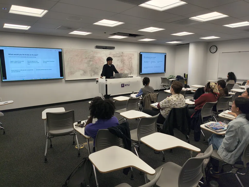 The classroom from the side: Siddharth Vohra speaking at the front between two screens showing the same slide, with students at their desks.