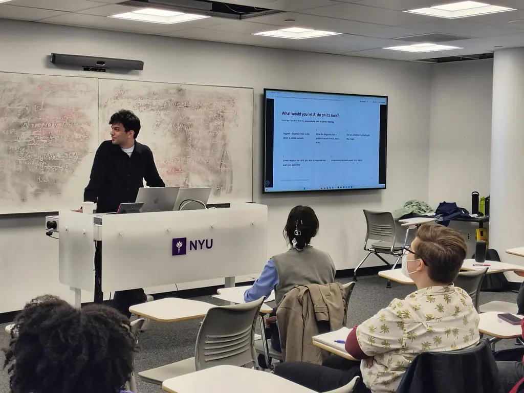 Siddharth Vohra at an NYU lectern, next to a screen showing the slide "What would you let AI do on its own?", with students seated in the foreground.